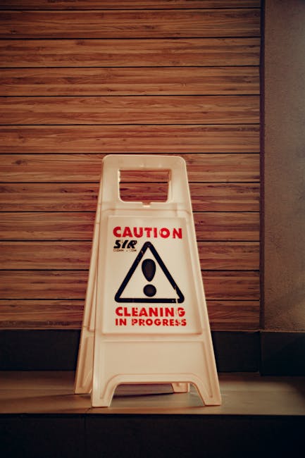 A light blue dustpan hanging on a plain white wall with a wooden-handled scrub brush resting inside it. The scrub brush has a curved wooden handle and beige bristles. The scene is well-lit with soft,
